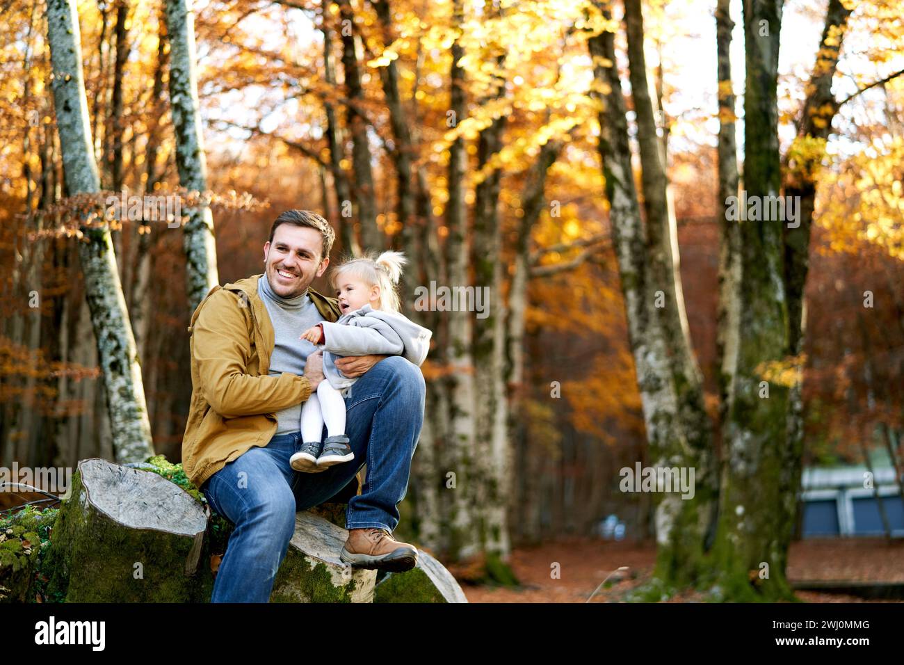 La bambina punta il padre in lontananza mentre si siede in ginocchio su un ceppo in un parco autunnale Foto Stock