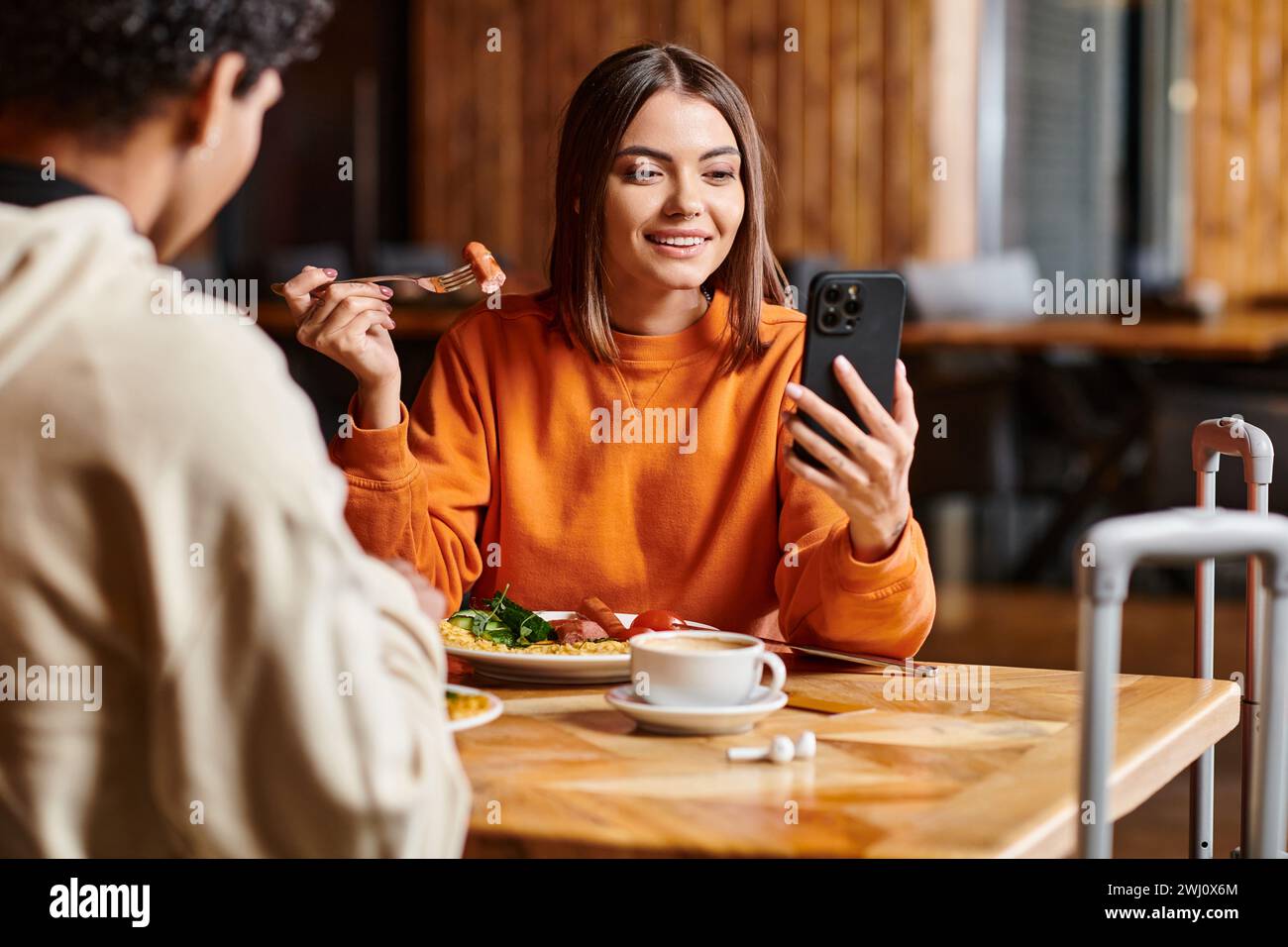Giovane donna in un maglione arancione vivace che guarda felicemente il suo telefono vicino al ragazzo nero durante il pasto Foto Stock