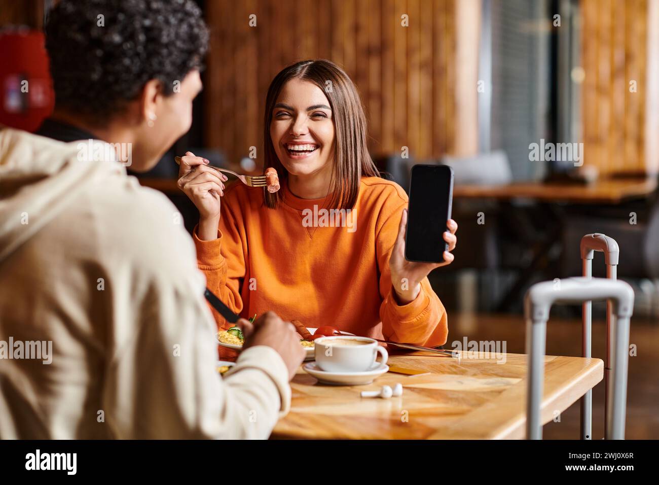 giovane donna felice con maglione arancione che mostra felicemente il suo telefono al ragazzo nero durante il pasto Foto Stock
