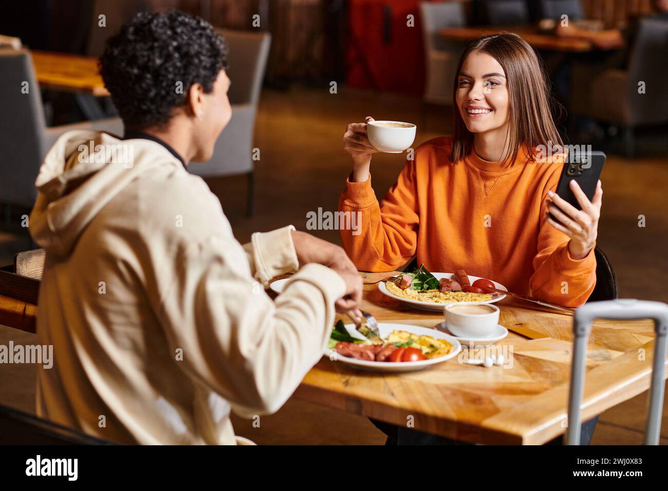 donna felice in maglione arancione che tiene il telefono e la tazza di caffè vicino al ragazzo nero durante il pasto Foto Stock