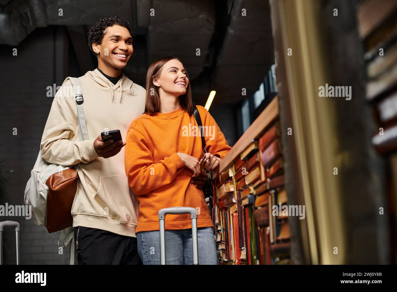 coppia felice e variegata con smartphone che condivide un momento di gioia in ostello vicino a una libreria Foto Stock