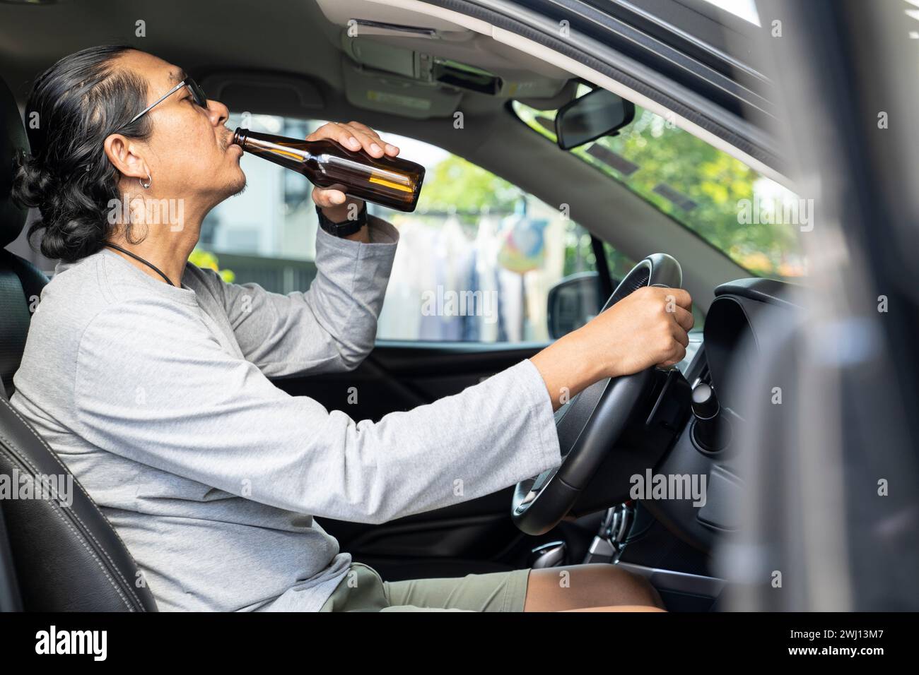 Un giovane asiatico ubriaco guida un'auto con una bottiglia di birra, un concetto di guida pericoloso. Foto Stock