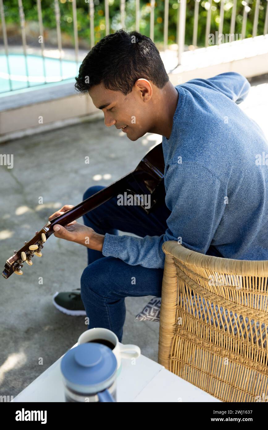 Felice uomo birazziale seduto sul balcone a suonare la chitarra a casa Foto Stock