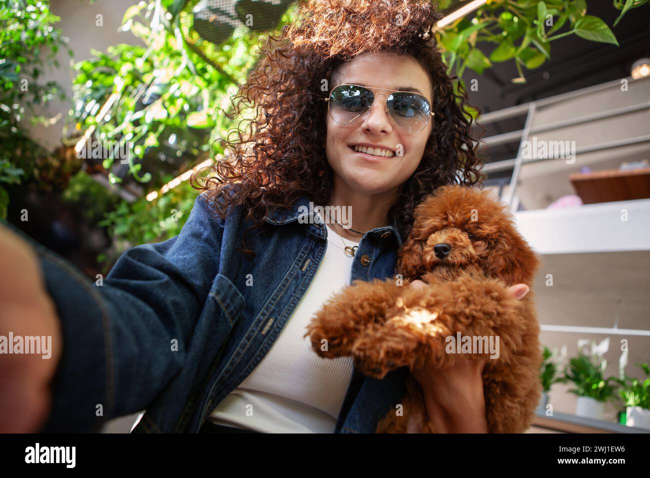 Selfie donna bruna ritratto con capelli ricci che tiene un piccolo cane marrone e sorride all'interno. Una vera donna felice con un animale domestico. Foto Stock