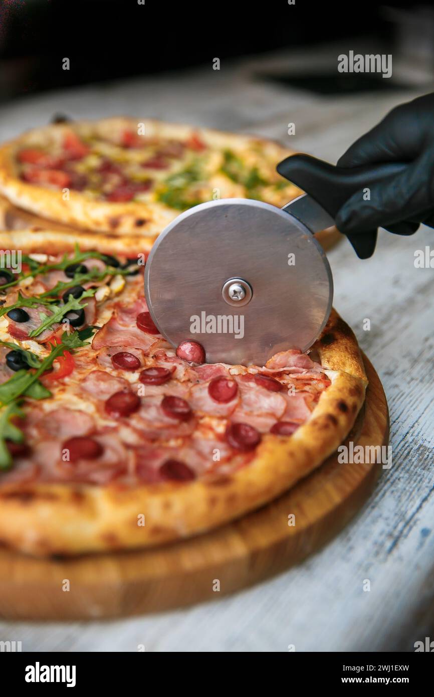 Tagliare la pizza a fette con un coltello speciale. Pizza fresca sullo sfondo. Coltello con le mani. Foto Stock