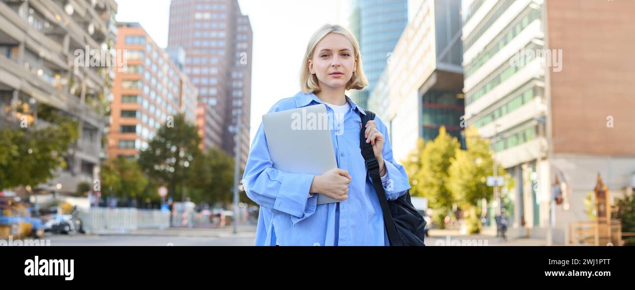 Ritratto di una giovane donna sicura di sé, studentessa universitaria con zaino e laptop, che va a lezione, in piedi all'aperto su una strada vuota Foto Stock