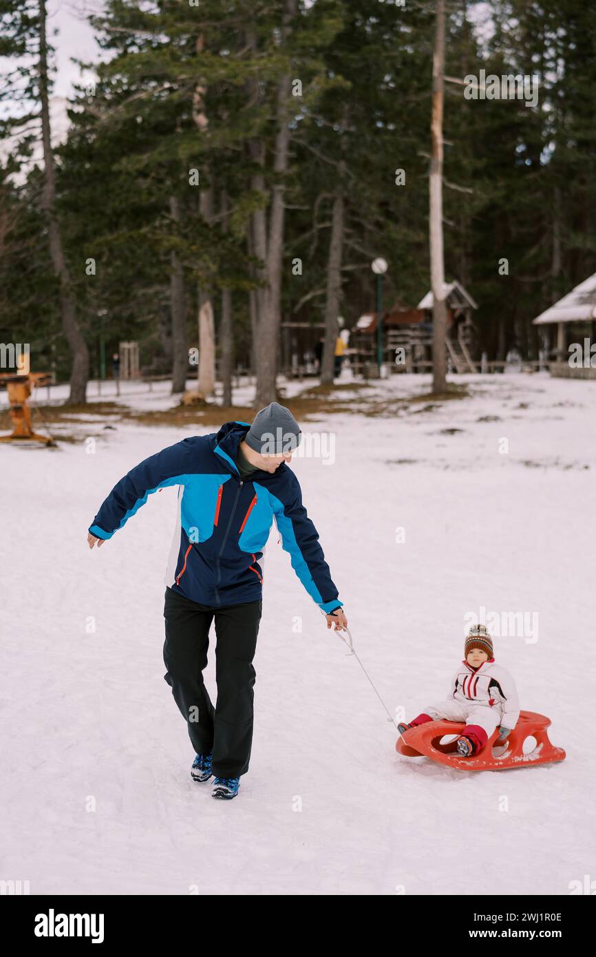 Papà porta un bambino piccolo su una slitta lungo il bordo innevato della foresta, guardando indietro Foto Stock