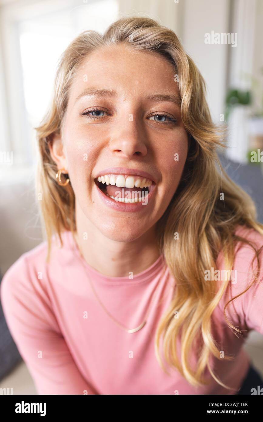 Felice bionda donna caucasica che sta videochiamando e sorridendo nel soleggiato soggiorno Foto Stock