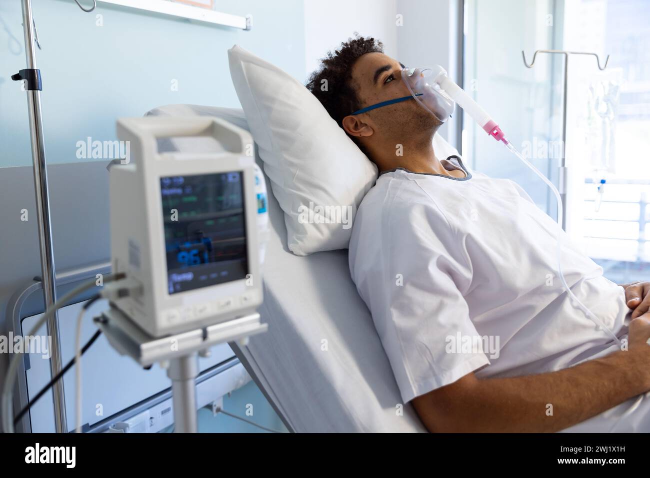 Paziente maschio birazziale con maschera di ossigeno sdraiato a letto in soleggiata stanza d'ospedale Foto Stock