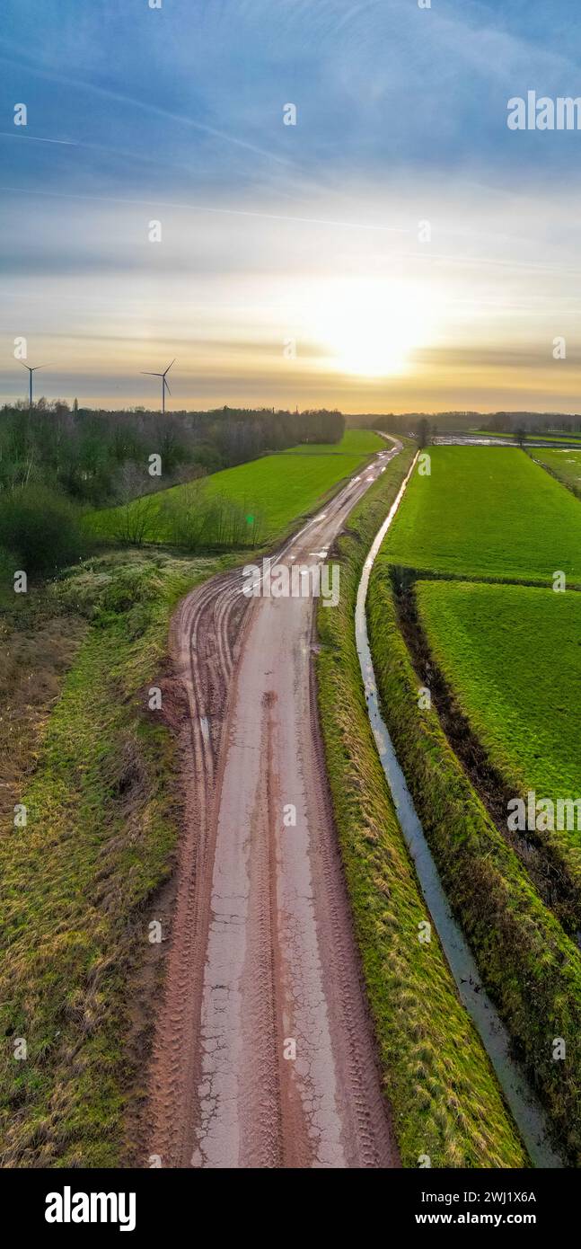 Questa immagine è una vista aerea che cattura le curve dolci di un sentiero rurale fangoso mentre si snoda attraverso i vibranti campi verdi. Il sentiero conduce verso l'orizzonte dove il sole del mattino presto irradia una luce soffusa e calda, suggerendo l'inizio di una nuova giornata. Il contrasto tra il sentiero fangoso e i campi lussureggianti è impressionante, sottolineando la bellezza del paesaggio rurale. Sullo sfondo, le turbine eoliche si allineano al sole nascente, simboleggiando un'armonia tra agricoltura ed energia rinnovabile. Ripresa aerea del Curvy Rural Path all'alba. Foto di alta qualità Foto Stock