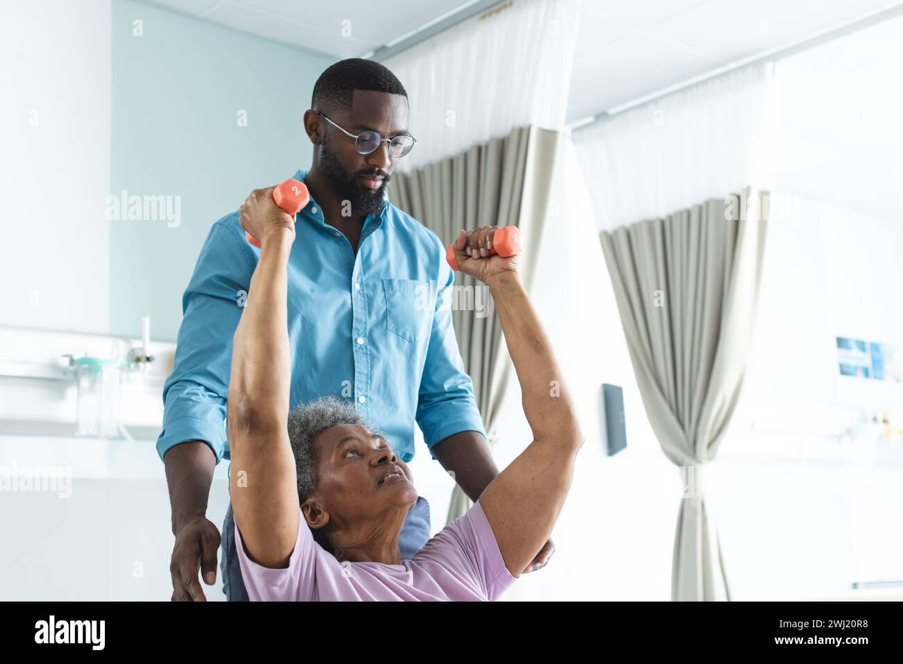 Donna anziana afroamericana che si esercita con pesi e medico maschile che consiglia in camera d'ospedale Foto Stock