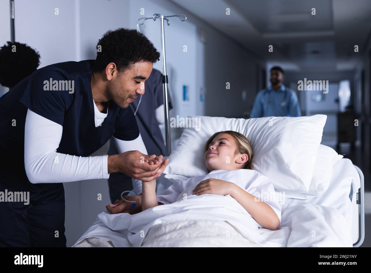 Medico maschile diversificato che parla con la ragazza sdraiata a letto nel corridoio dell'ospedale Foto Stock