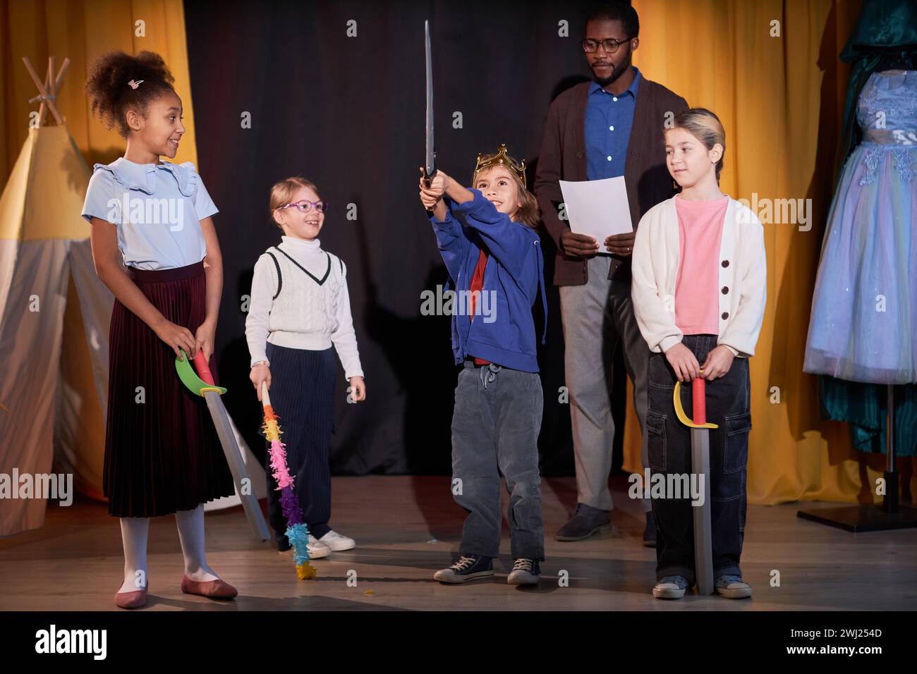 Ritratto a figura intera di un ragazzo che gioca a un valoroso principe sul palco a scuola con un gruppo di bambini Foto Stock