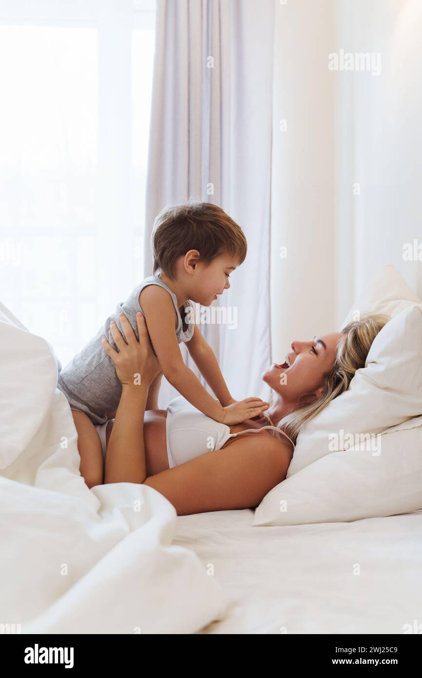 La madre felice e il figlio piccolo si impegnano in un gioco allegro, creando ricordi preziosi mentre si legano nel comfort del loro letto Foto Stock