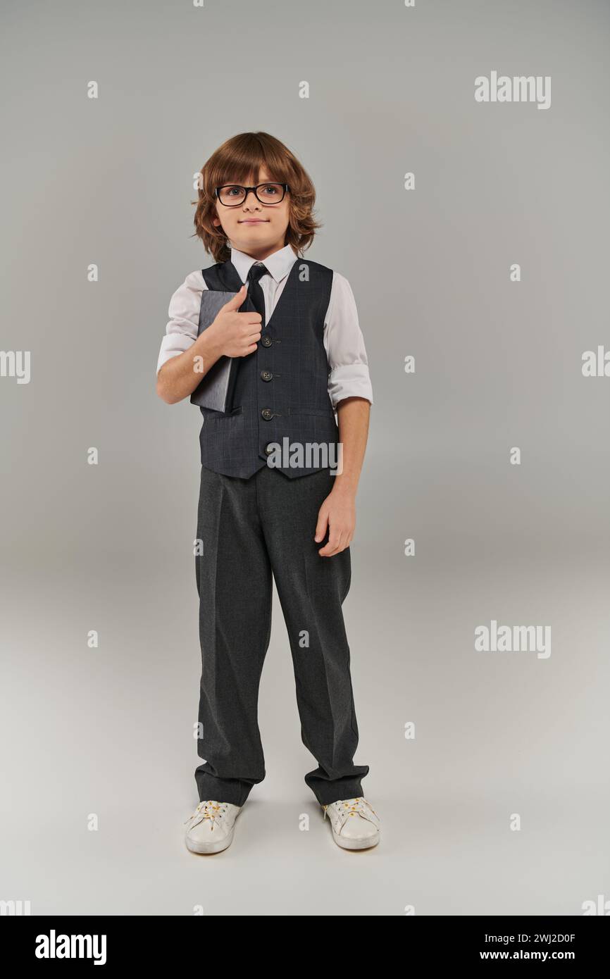 Un ragazzino vestito con un gilet alla moda e cravatta che tiene in mano un libro e sta in piedi su sfondo grigio Foto Stock