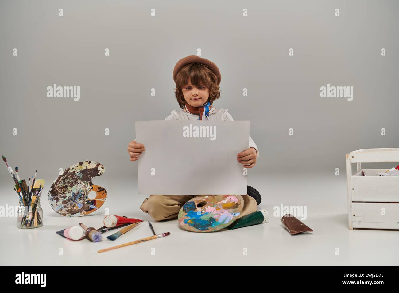 giovane artista circondato da colori vivaci e strumenti per dipingere, ragazzo che guarda la carta bianca Foto Stock