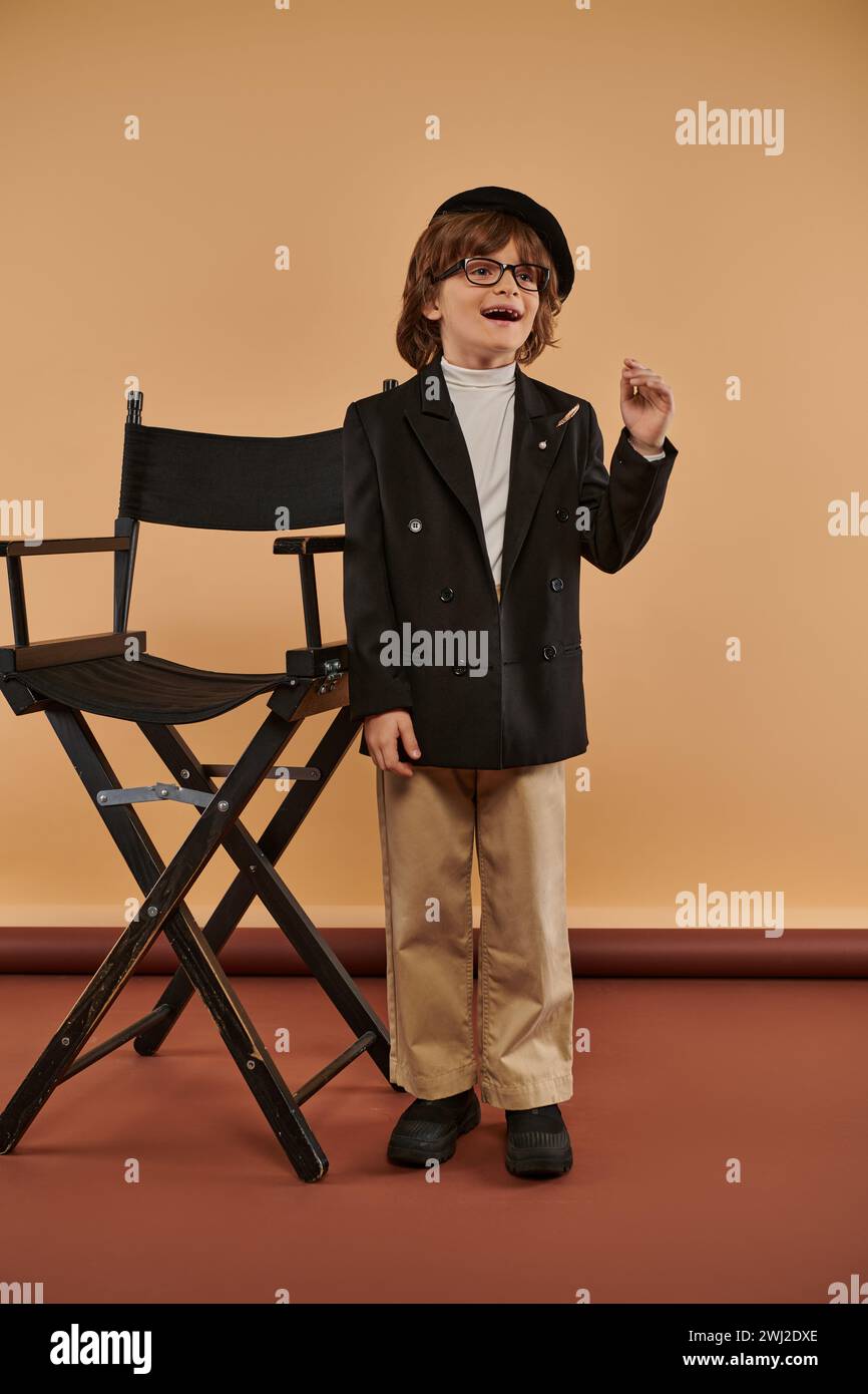 ragazzo ispirato in piedi vicino alla sedia del regista, con un sorriso sul viso che mostra il segno dell'idea, professione Foto Stock