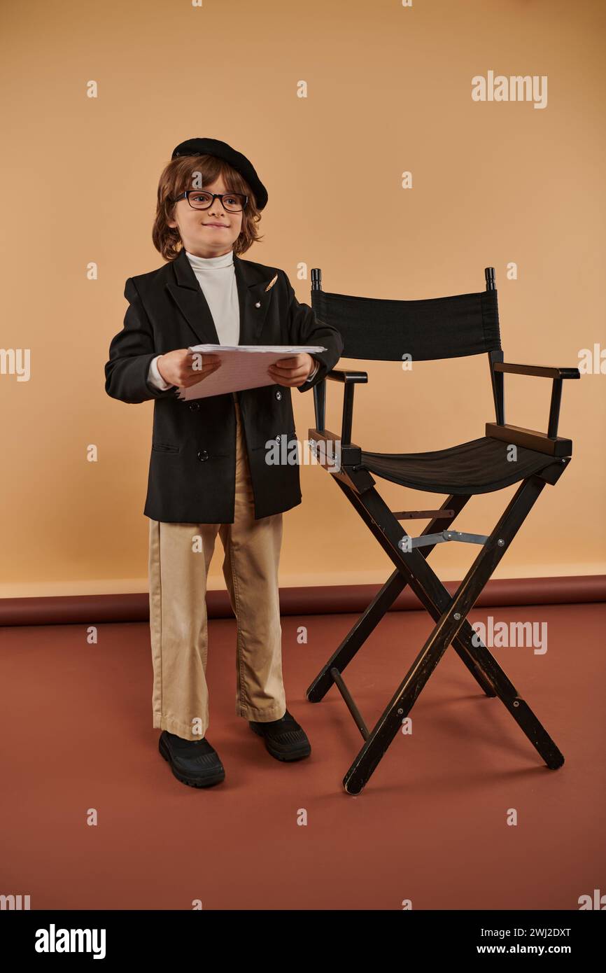 happy boy è orgoglioso di stare accanto alla sedia director, vestito con giacca e pantaloni, con i documenti in mano Foto Stock