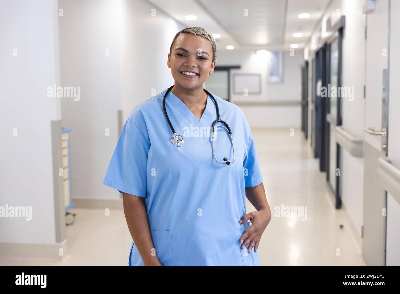 Ritratto di felice medico donna birazziale che indossa scrub e stetoscopio in corridoio in ospedale Foto Stock