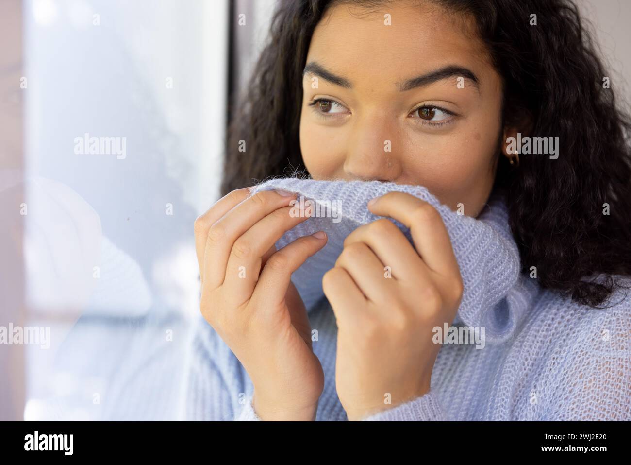 Donna birazziale che si copre di maglione e guarda fuori dalla finestra di casa soleggiata Foto Stock
