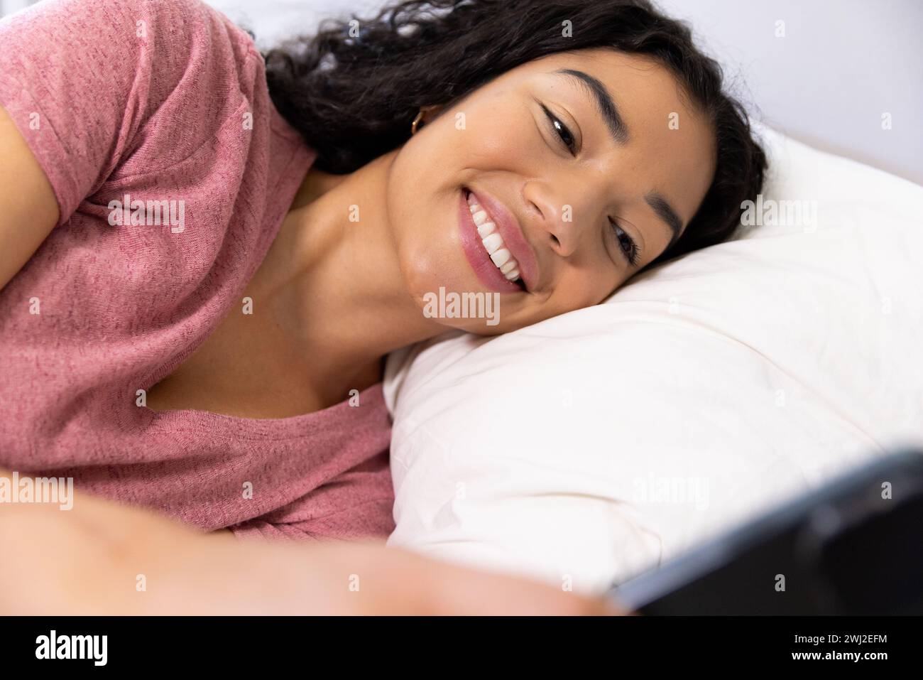 Felice donna birazziale che usa lo smartphone sdraiato sul letto a casa soleggiata Foto Stock