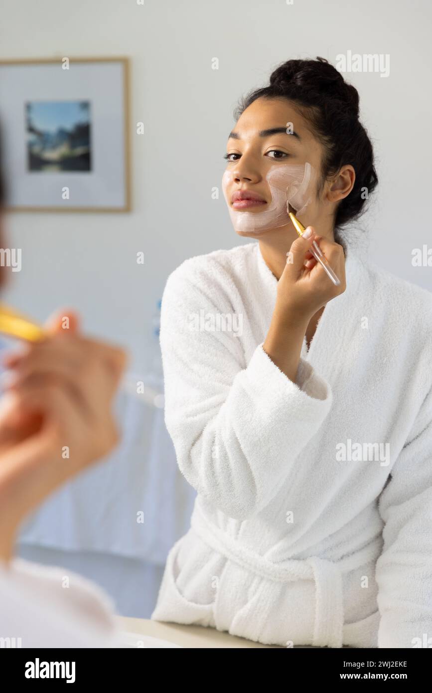 Donna birazziale in accappatoio applicazione maschera utilizzando pennello in bagno soleggiato Foto Stock