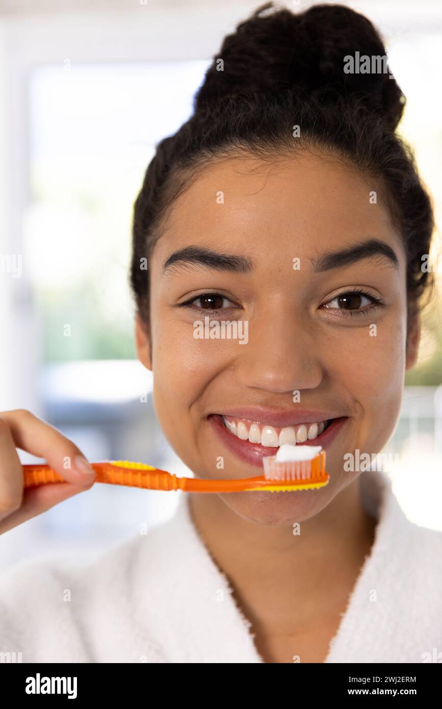 Ritratto di una donna birazziale felice in accappatoio che regge una spazzola con pasta dentale nel bagno soleggiato Foto Stock