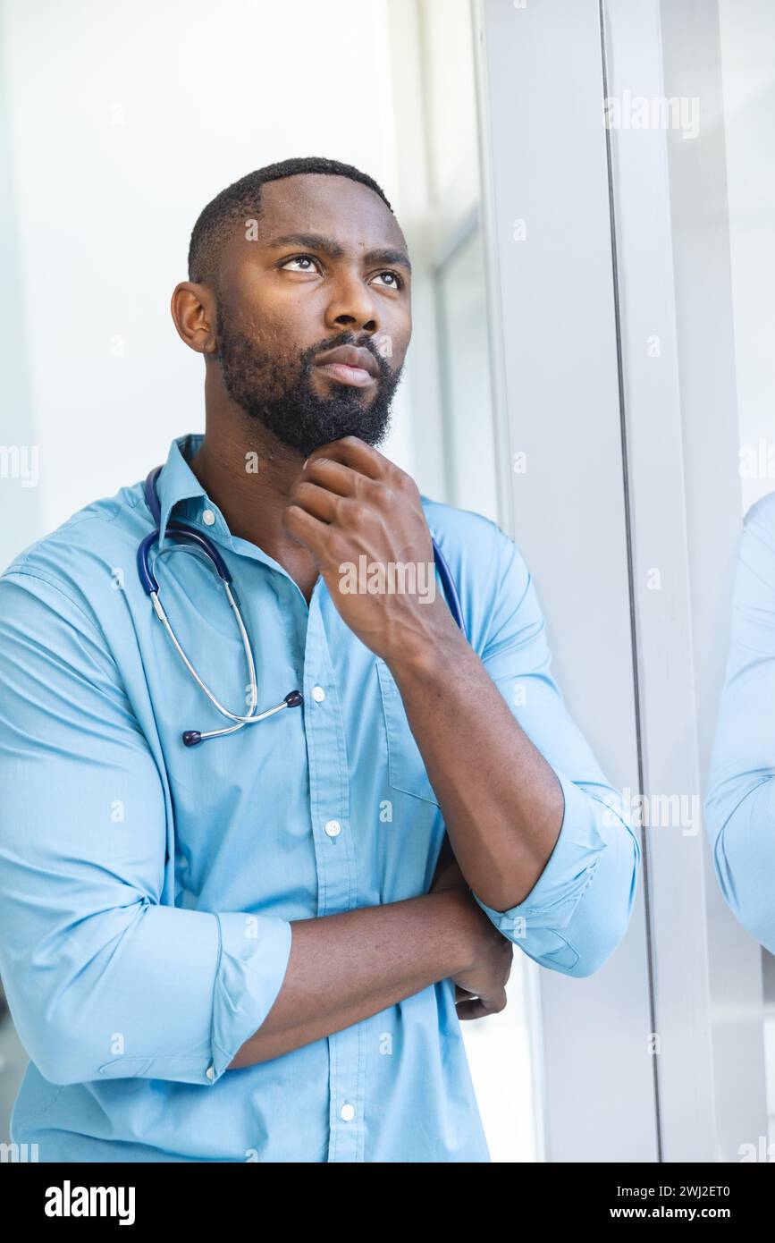 Dottore africano americano premuroso che indossa la camicia blu guardando attraverso la finestra nella stanza d'ospedale Foto Stock
