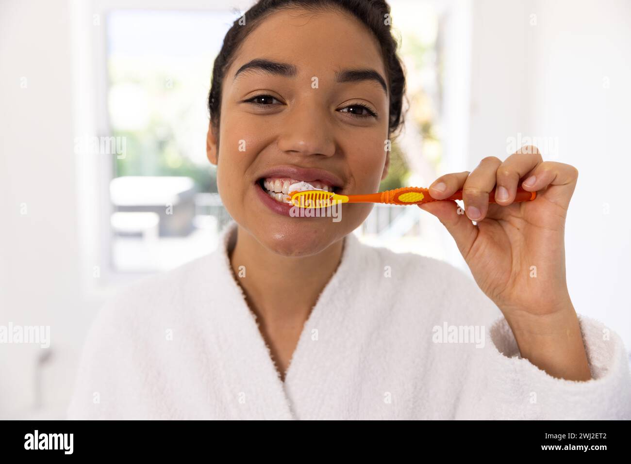 Ritratto di donna birazziale in accappatoio che spazzola i denti nel bagno soleggiato Foto Stock
