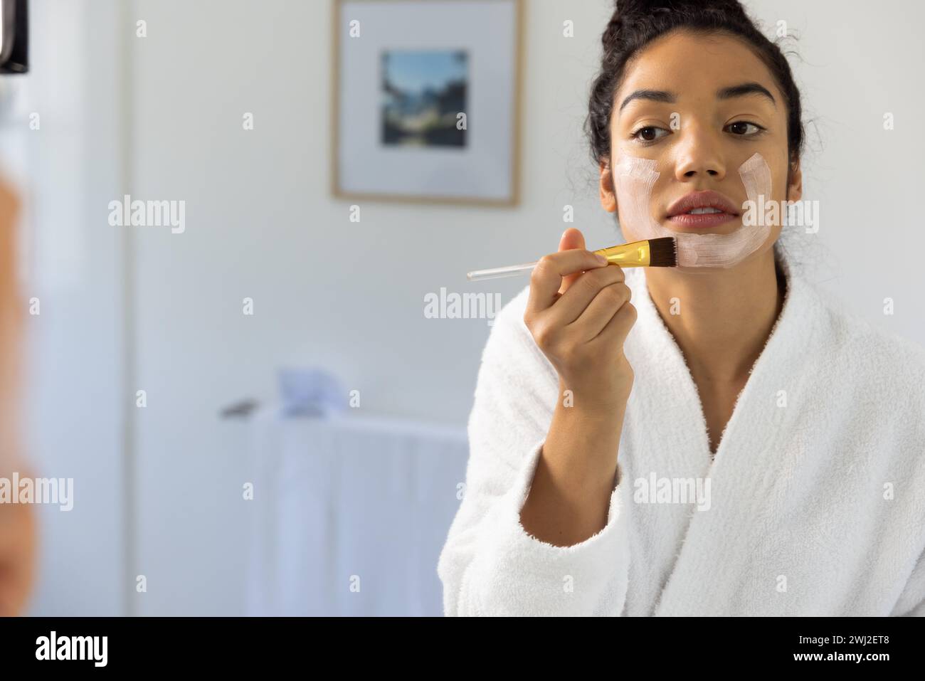 Donna birazziale in accappatoio applicazione maschera utilizzando pennello in bagno soleggiato Foto Stock