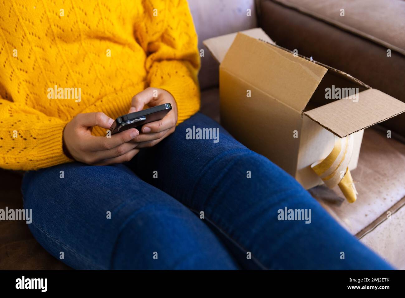 Sezione centrale della donna birazziale in maglione giallo con pacchetto che utilizza lo smartphone a casa Foto Stock