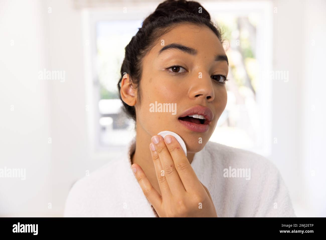 Ritratto di donna birazziale in accappatoio che pulisce il viso in un bagno soleggiato Foto Stock