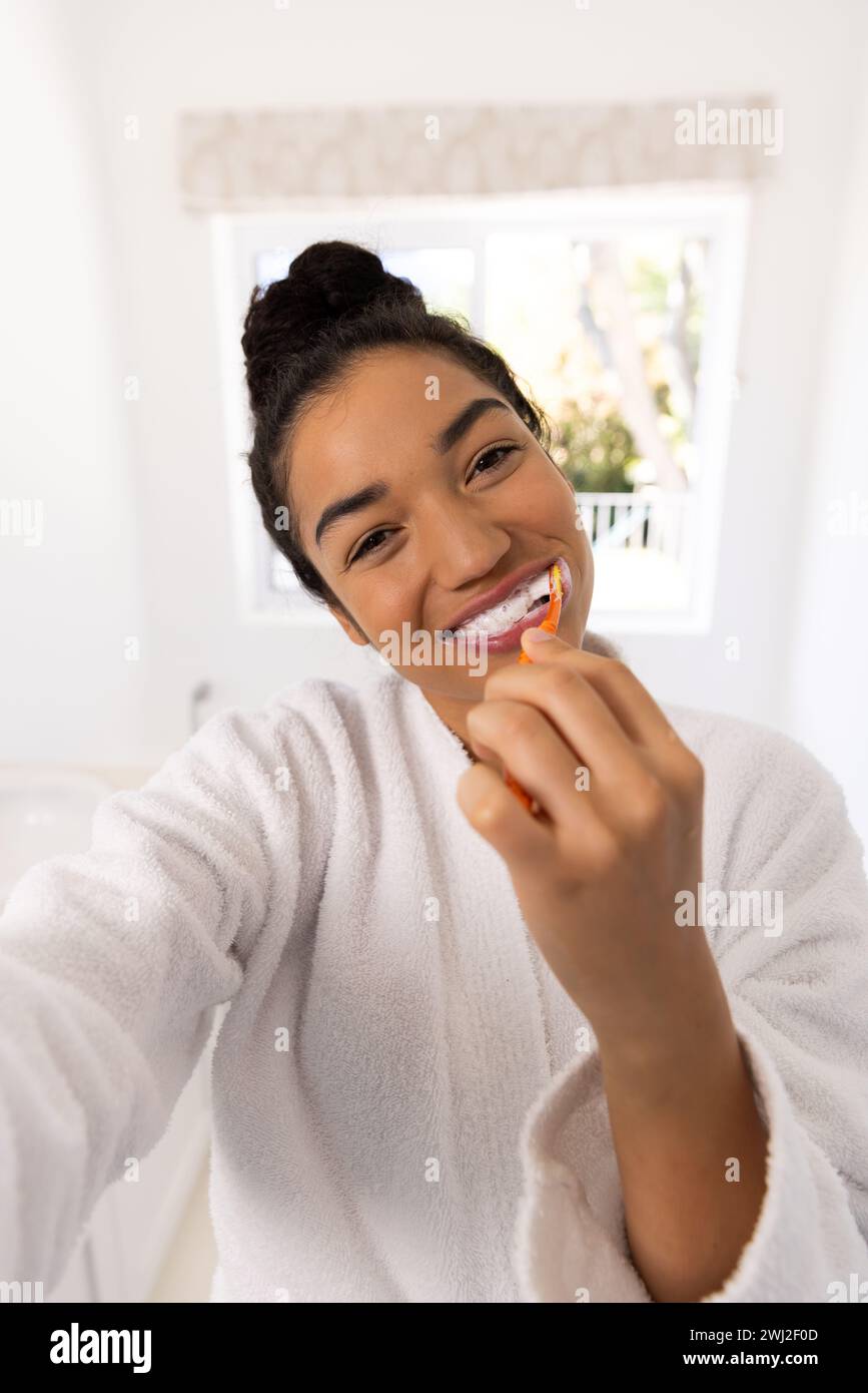 Ritratto di una donna birazziale felice in accappatoio che spazzola i denti nel bagno soleggiato Foto Stock