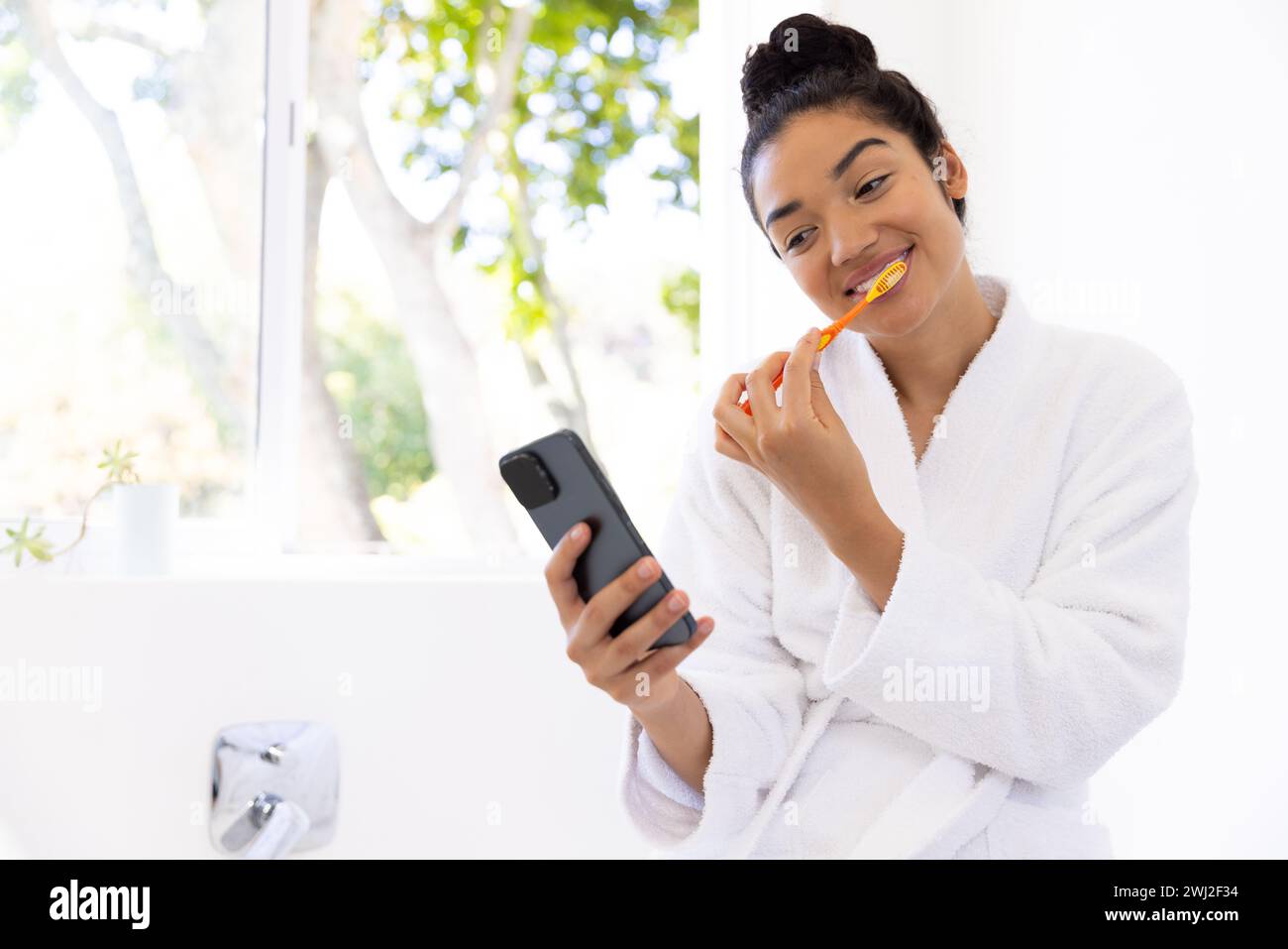 Donna birazziale in accappatoio che pulisce i denti e utilizza lo smartphone nel bagno soleggiato Foto Stock