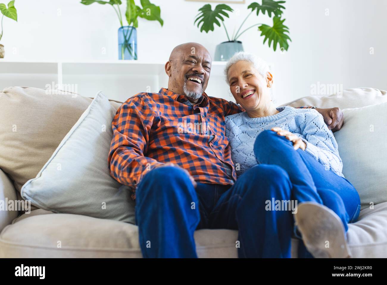 Coppie anziane felici e diverse sedute sul divano e abbracciate nel soggiorno soleggiato Foto Stock