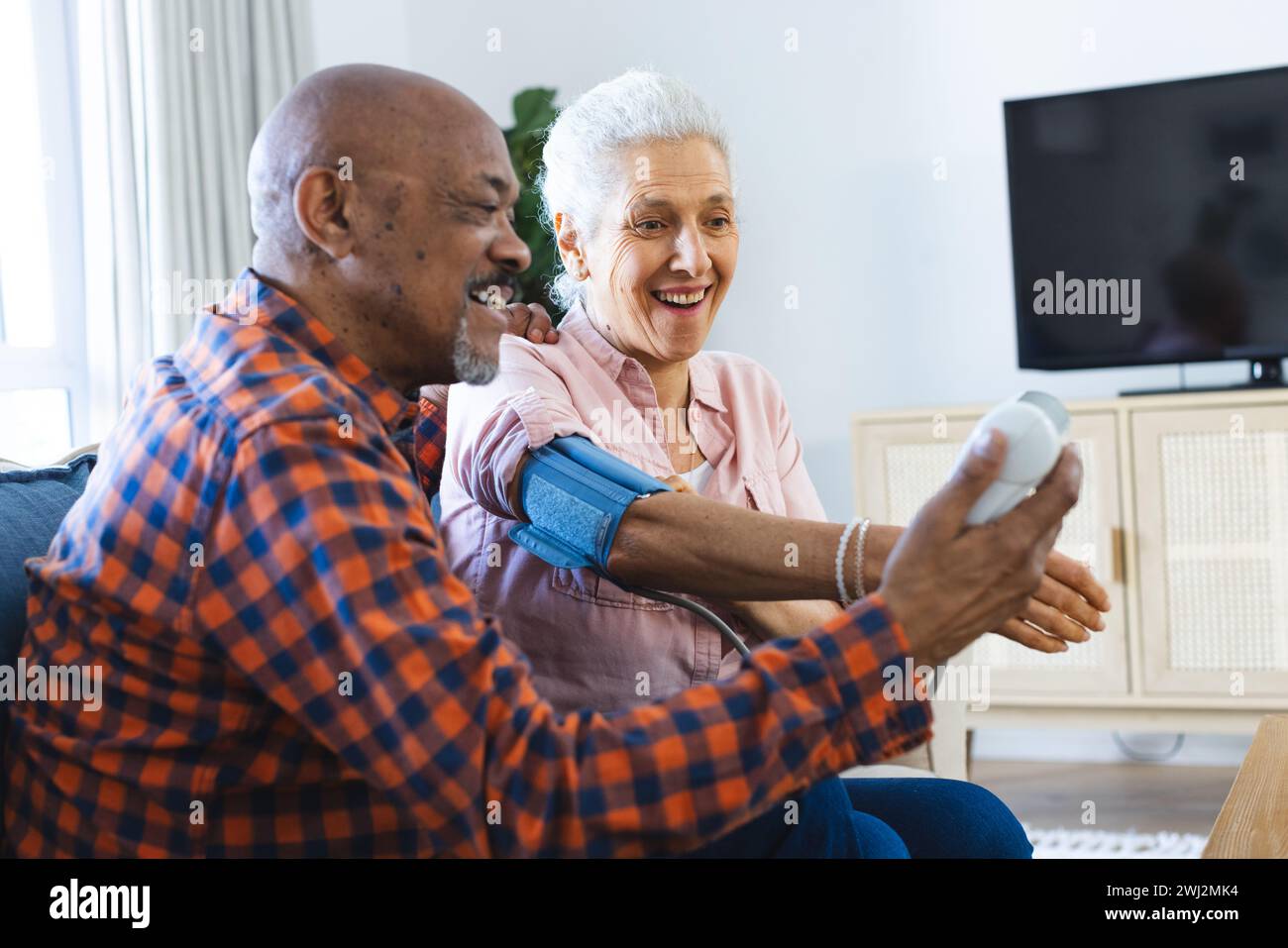 Felice e diversificato uomo anziano che controlla la pressione sanguigna di una donna anziana in un soggiorno soleggiato Foto Stock