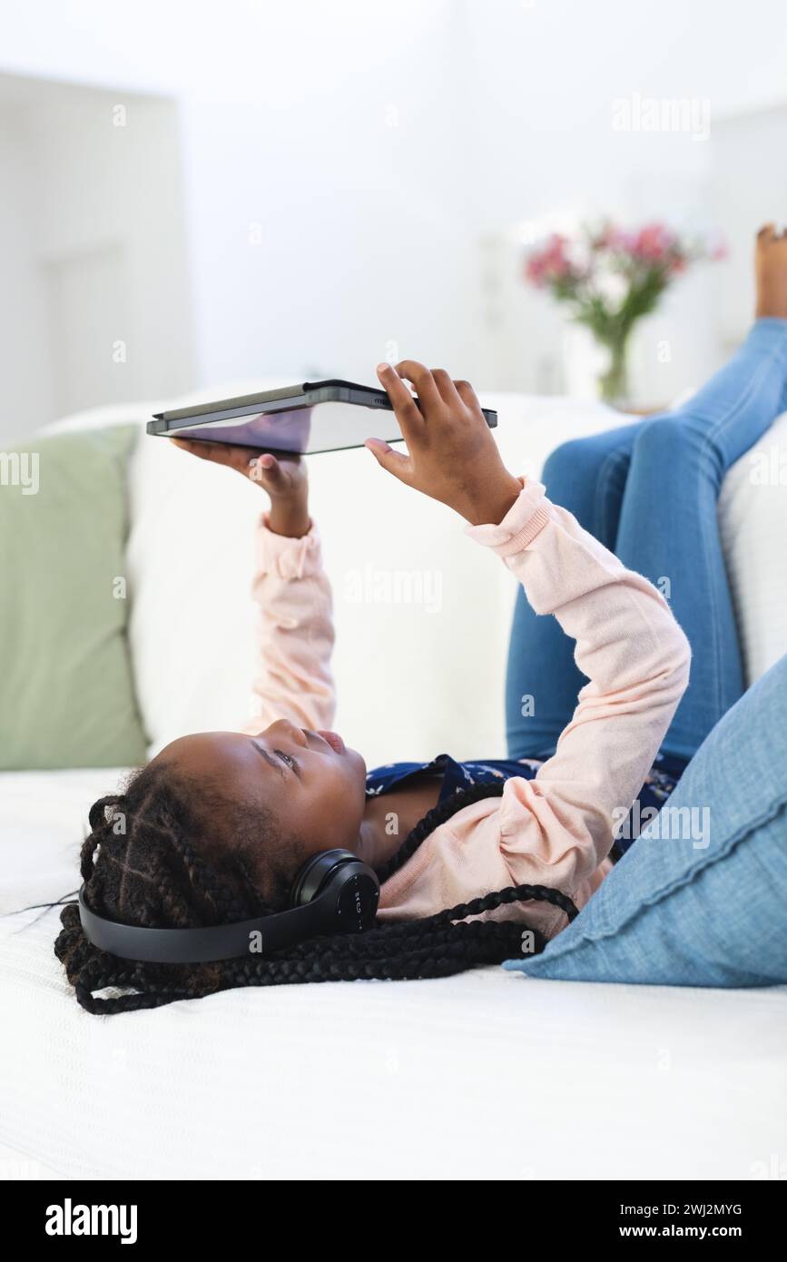 Felice ragazza afroamericana sdraiata sul divano, ascoltando musica e utilizzando un tablet a casa, copiando spazio Foto Stock