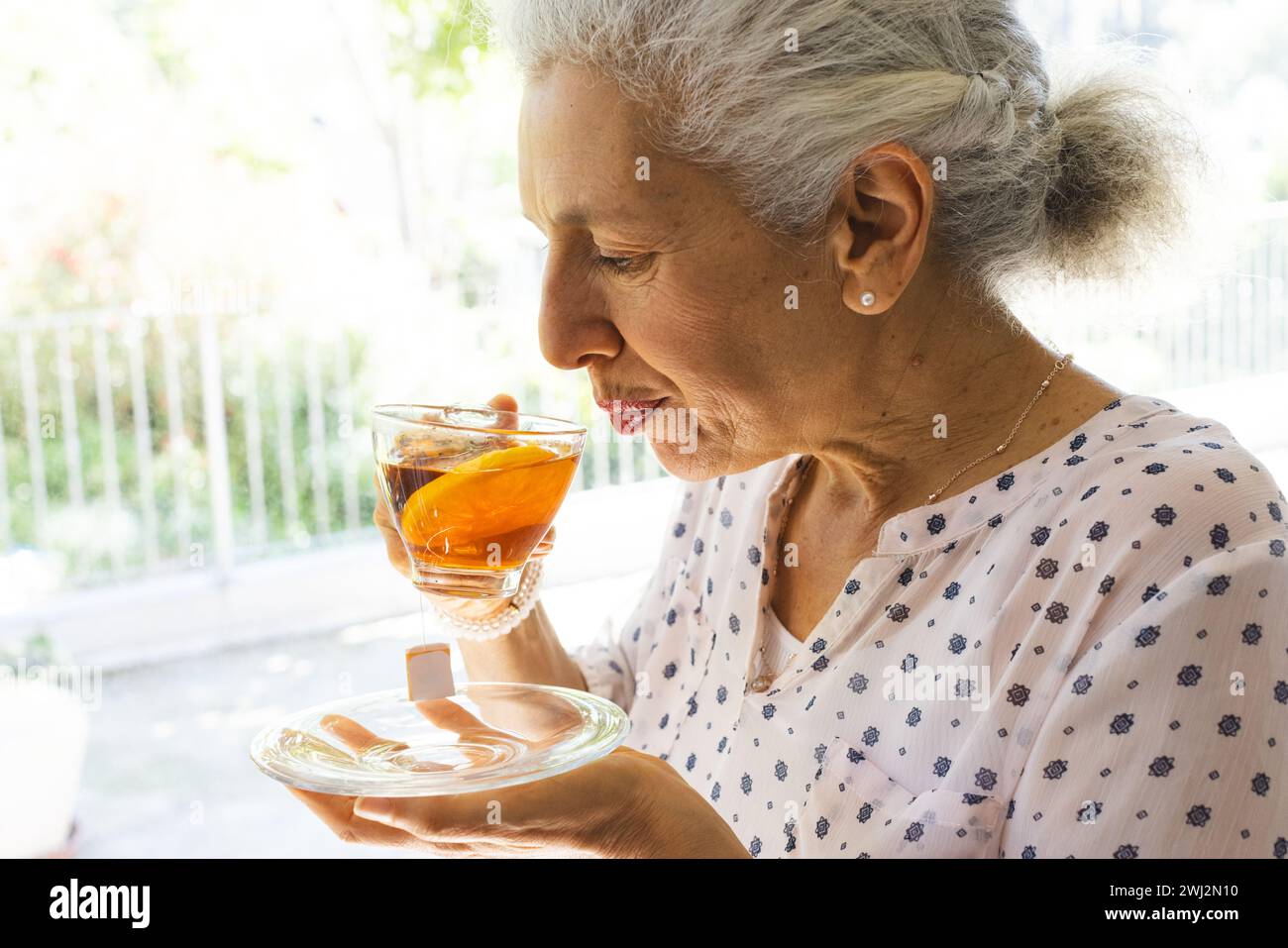 Donna anziana caucasica che beve il tè alla finestra di una casa soleggiata Foto Stock