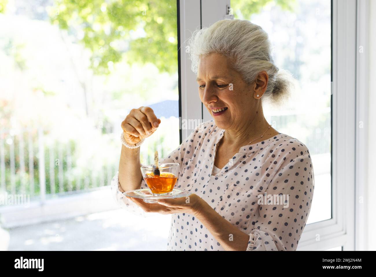 Felice donna anziana caucasica che beve il tè alla finestra nella casa soleggiata Foto Stock