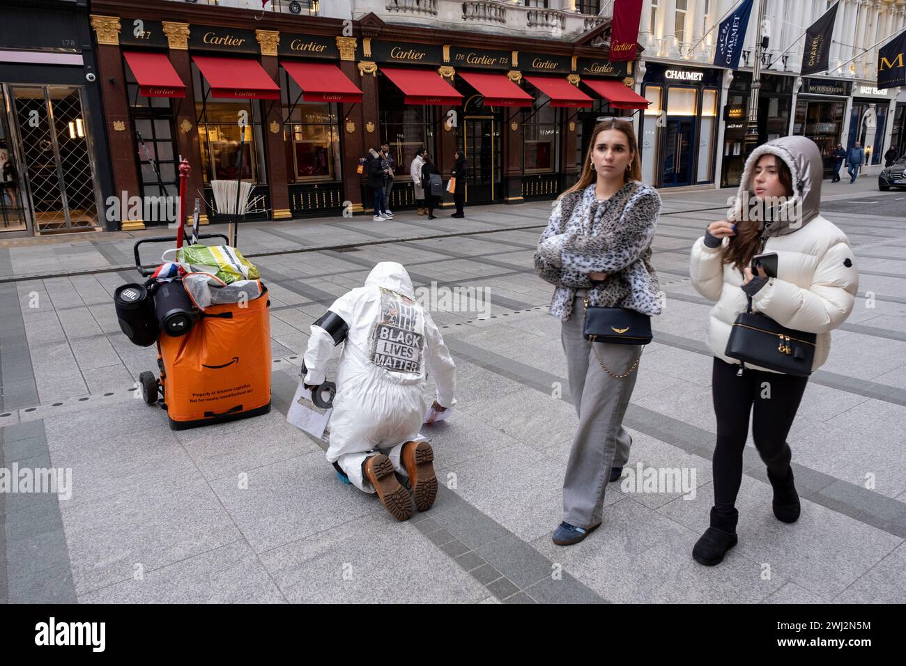 La gente passa davanti a un uomo in Bond Street che indossa tute bianche e un cartello Black Lives Matter attacca lettere giù sul marciapiede in una protesta con un messaggio poco chiaro il 4 febbraio 2024 a Londra, Regno Unito. Foto Stock