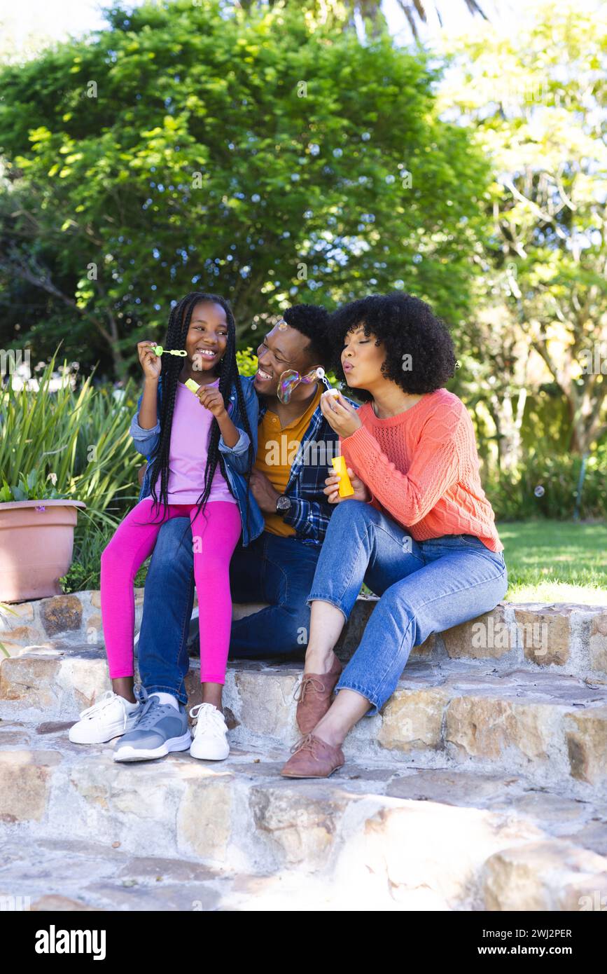 Felice padre, madre e figlia afroamericani che soffiano bolle nel giardino soleggiato, spazio copia Foto Stock