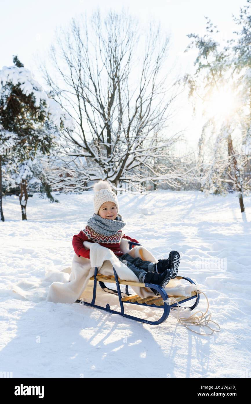 Bambino seduto sulla slitta in un parco cittadino innevato durante le soleggiate giornate invernali Foto Stock