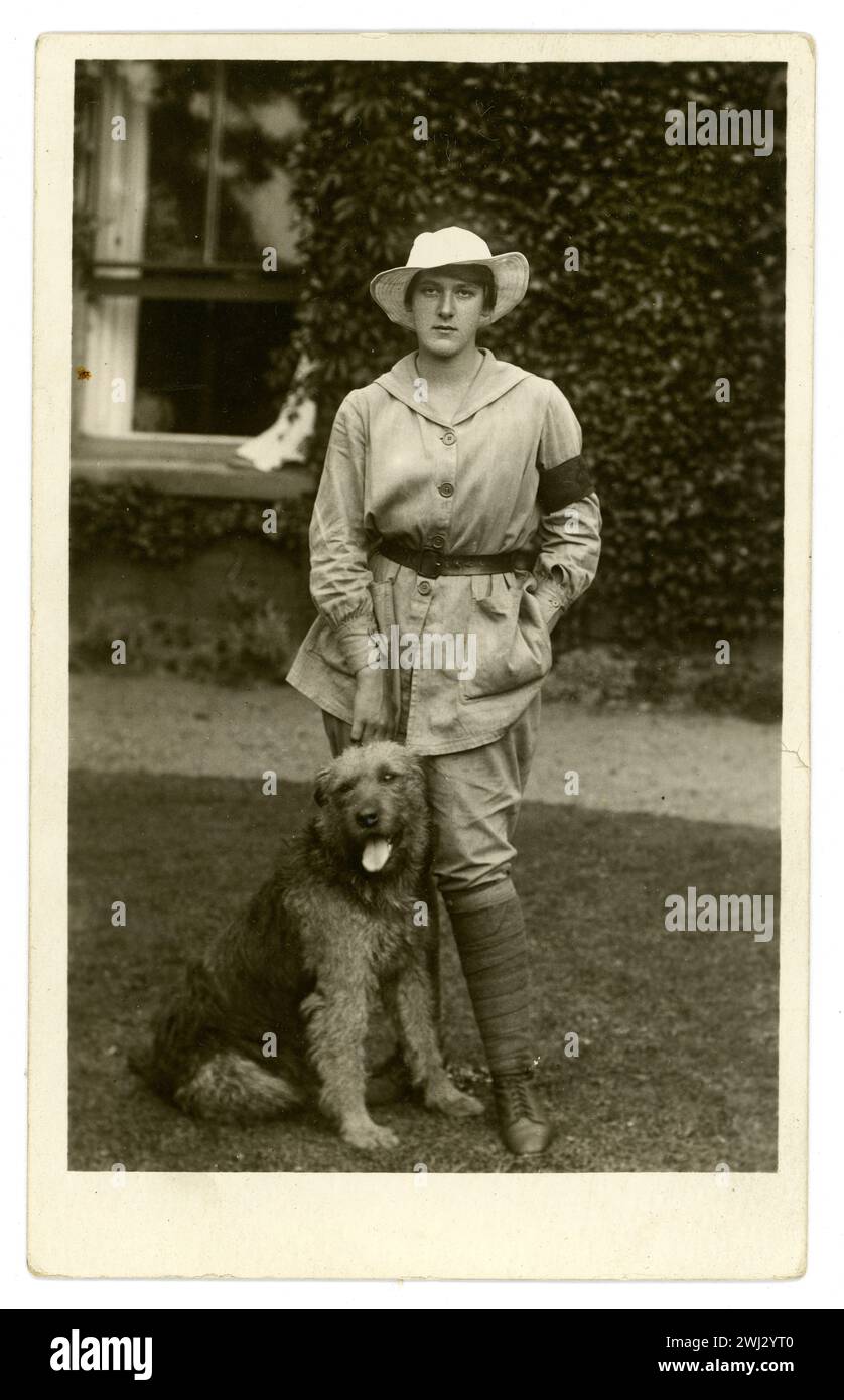 Cartolina originale dell'era WW1 di una ragazza di casa chiamata Esme Greene che indossa una tunica beige che denota i doveri generali della fattoria (al contrario dell'uniforme bianca per il volo) e una fascia da braccio di servizio di 3 mesi. Indossa i breeches, una tunica totale al ginocchio (con cintura). Si trova fuori da una casa di campagna o fattoria, con un cane chiamato Kim, datato 1917, Easton a Gordano, vicino a Bristol, Regno Unito Foto Stock