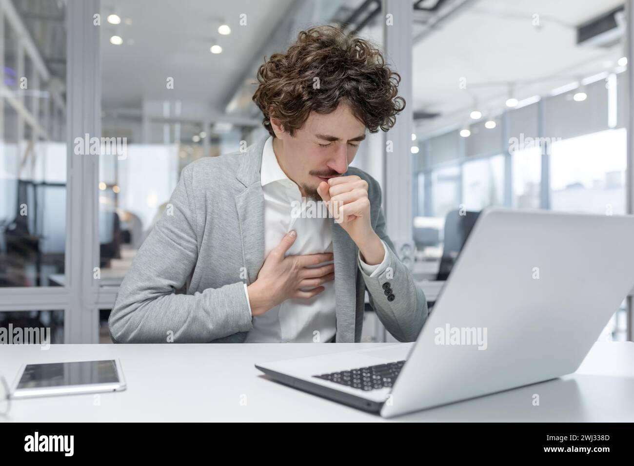 Lavoratore d'ufficio malato, giovane uomo d'affari seduto alla scrivania con un portatile e documenti e tosse, copre la bocca con la mano e tiene il petto, sente respiro corto e dolore. Foto Stock