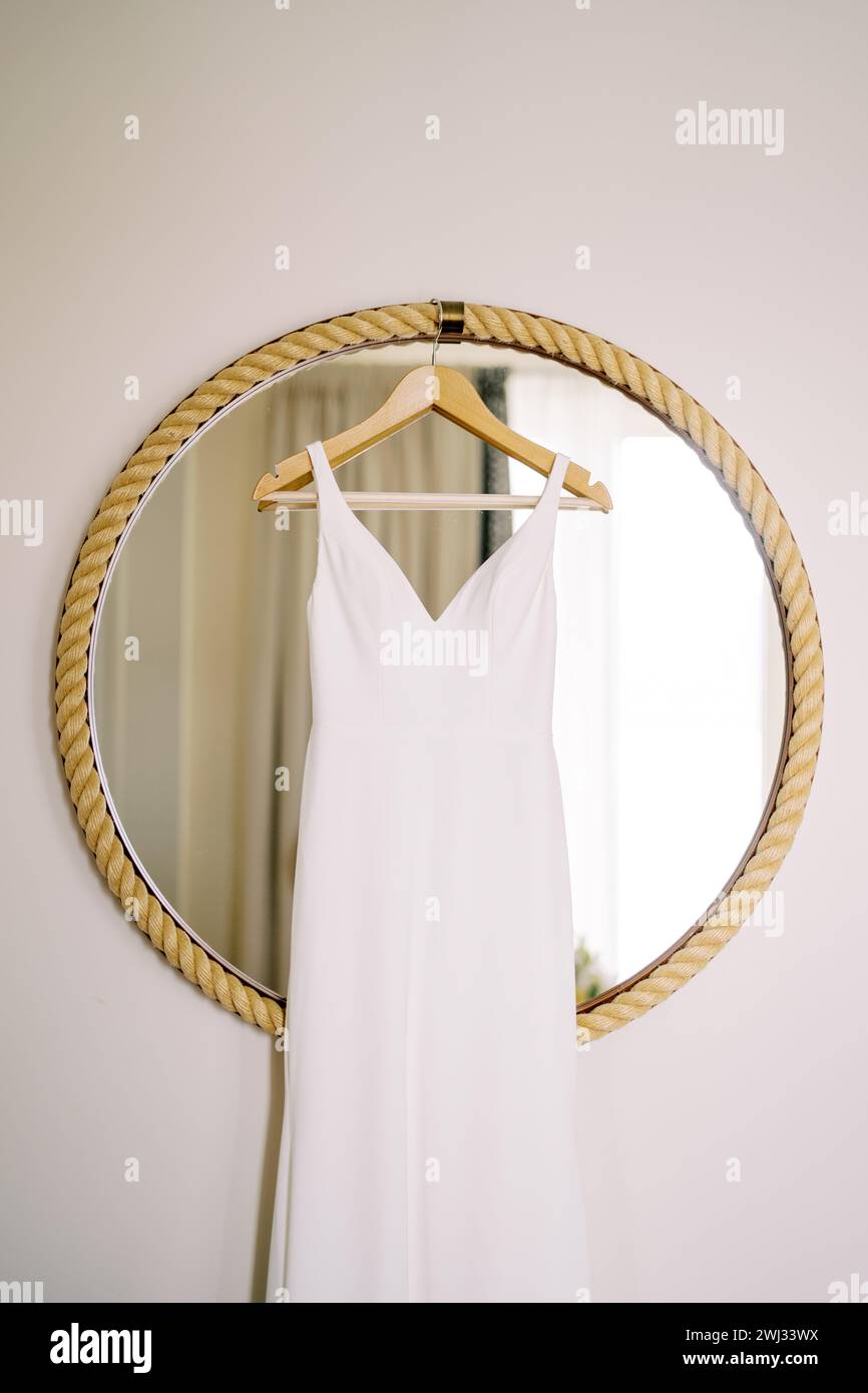 L'abito da sposa è appeso a un appendiabiti su uno specchio rotondo in una struttura a corda sulla parete della stanza Foto Stock