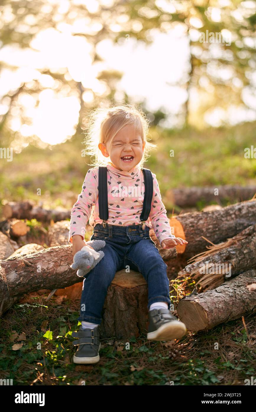 La bambina siede su un ceppo nella foresta con un giocattolo in mano e ride con gli occhi chiusi Foto Stock