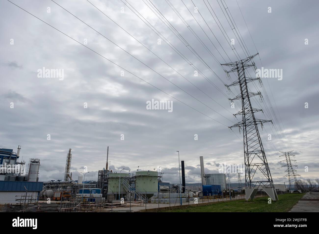 una torre elettrica di una linea ad alta tensione in un'area industriale con una fabbrica con camini, serbatoi e silos, in una giornata nuvolosa, ambiente industriale Foto Stock