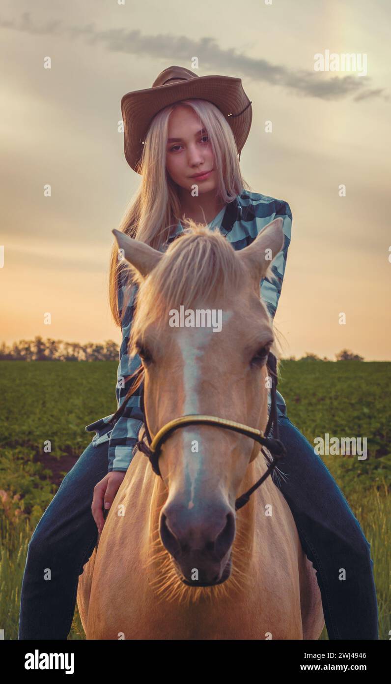 La ragazza vestita con un cappello da cowboy e jeans blu si siede su un cavallo sullo sfondo del tramonto sul ranch Foto Stock