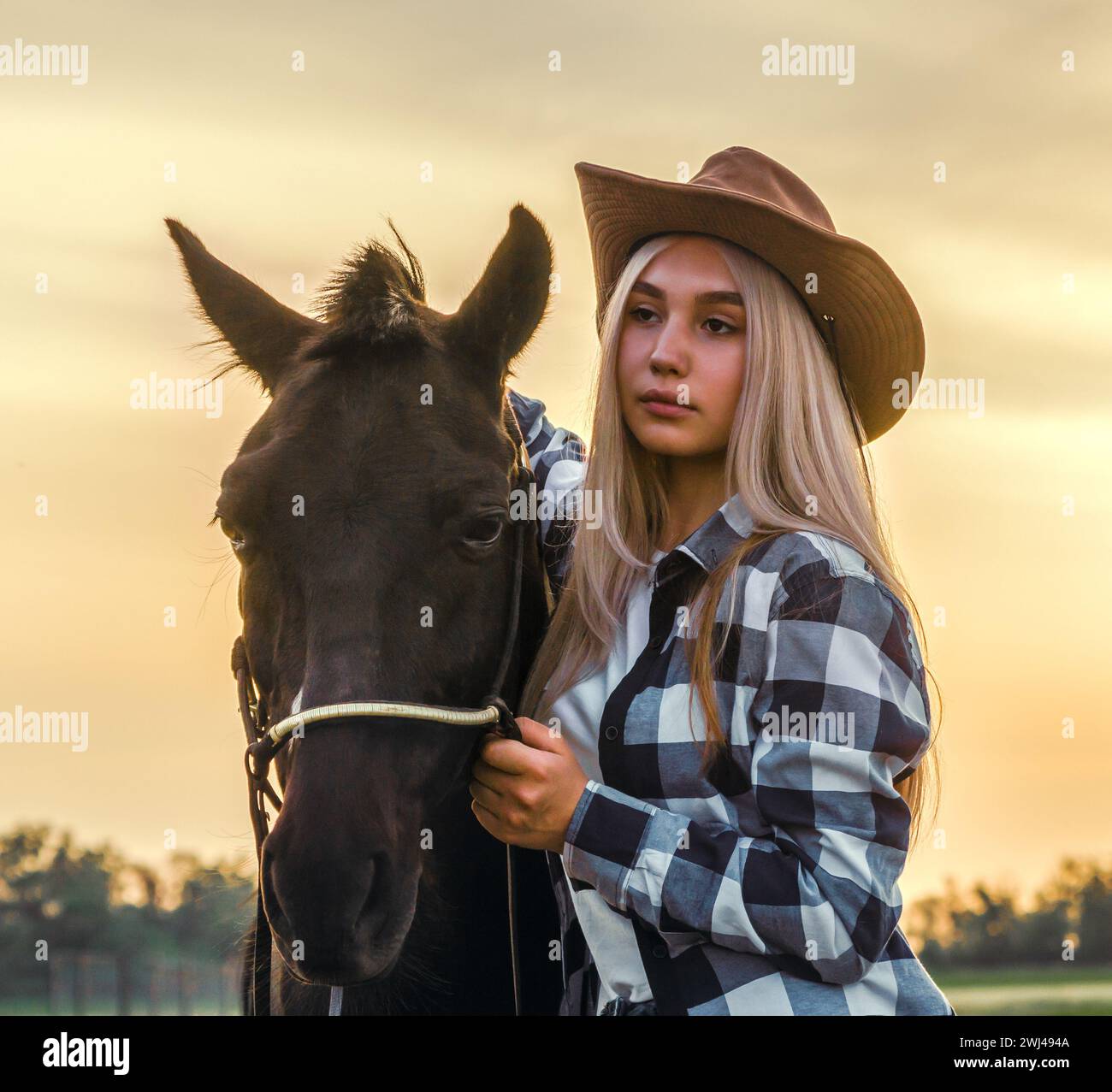 Giovane ragazza vestita con un cappello da cowboy e una camicia a quadri con un cavallo al tramonto al ranch Foto Stock