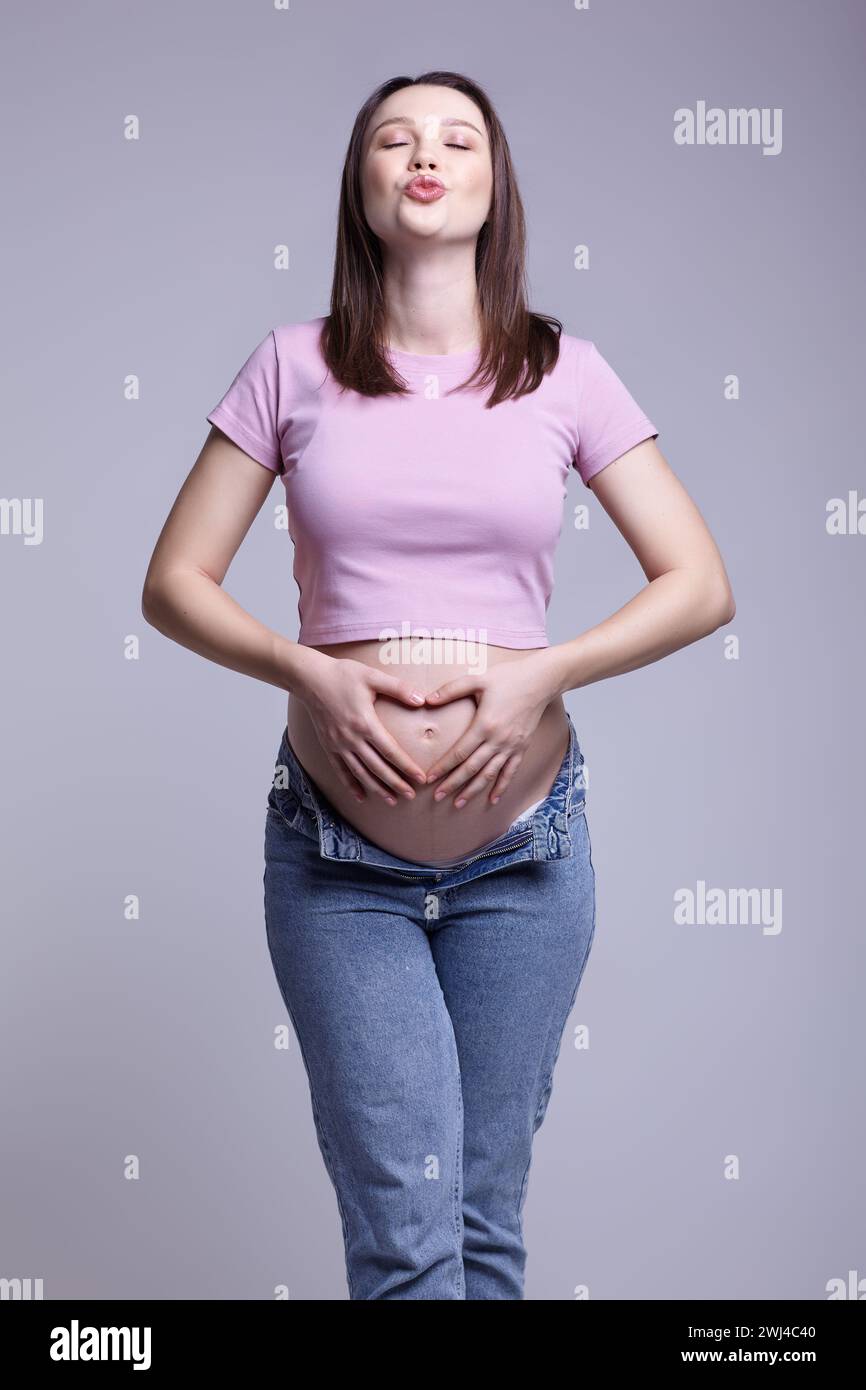 Giovane donna incinta con t-shirt rosa e jeans su sfondo grigio. Mani vicino alla pancia incinta con segno cardiaco. Labbra in un Foto Stock